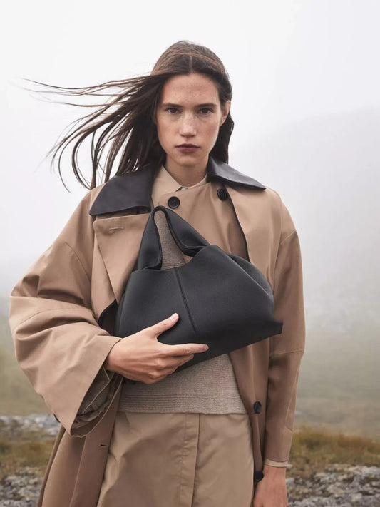 Woman in beige trench coat holding black leather handbag with foggy outdoor background