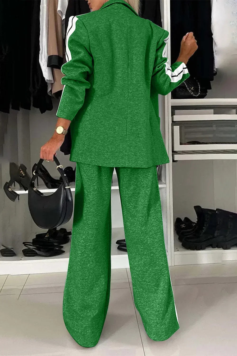 Woman wearing green wide-leg pants and matching jacket with white stripes, holding black handbag in stylish closet