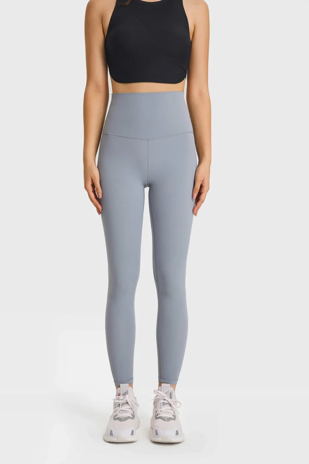 Woman wearing high-waisted light blue leggings and black crop top with white sneakers on plain background