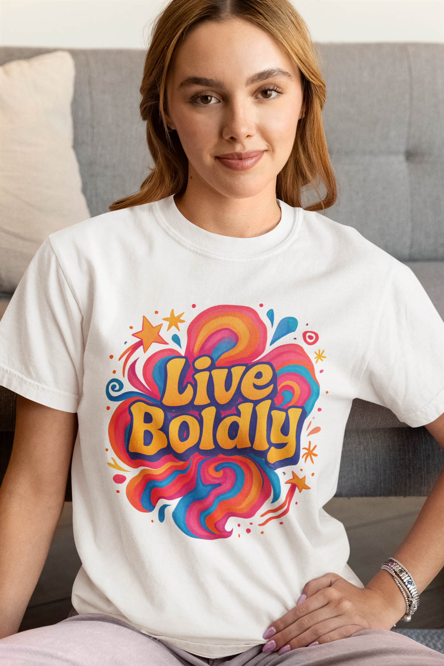 Woman wearing a white t-shirt with 'Live Boldly' design sitting on a couch.