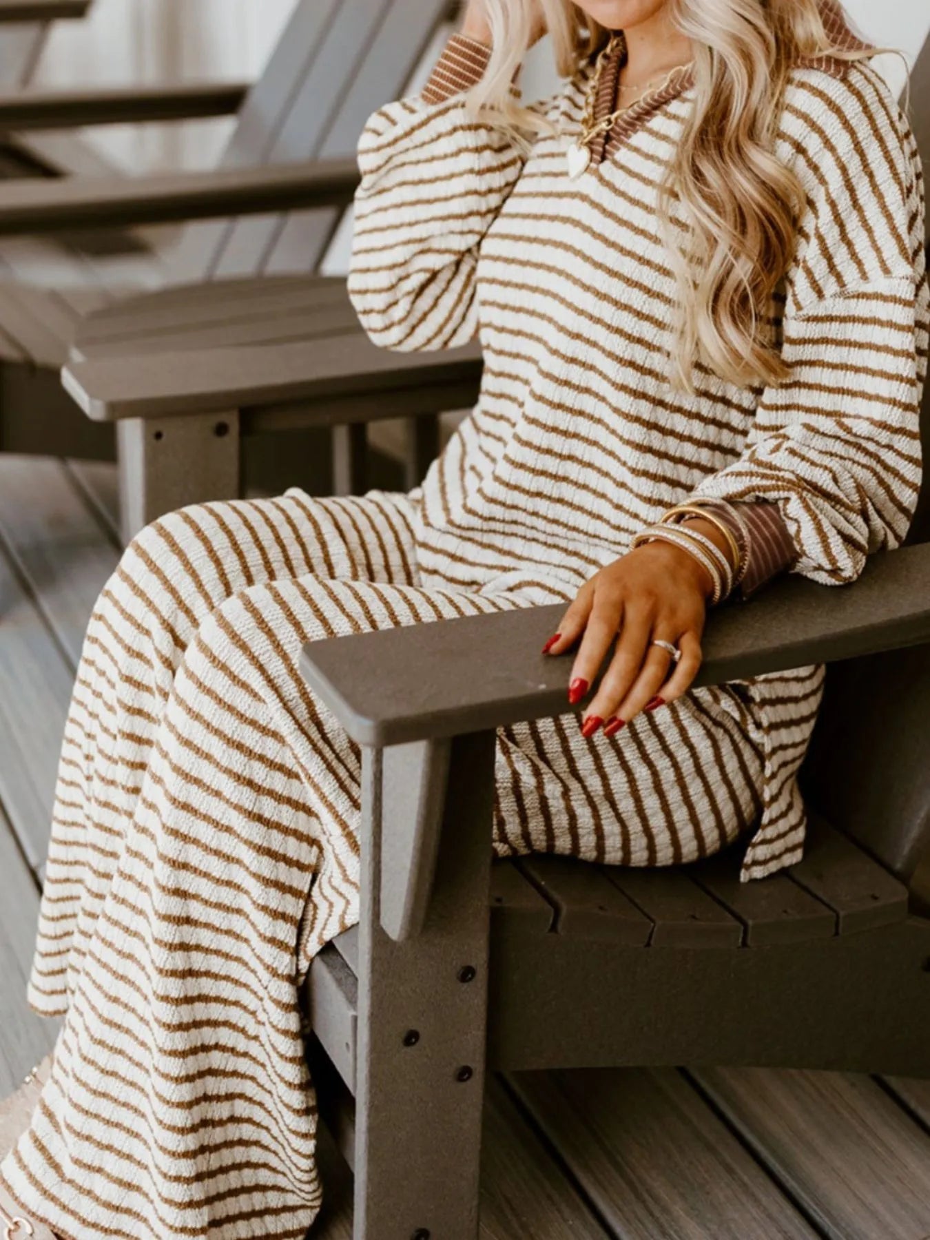 Woman in cozy beige and brown striped long sweater dress sitting on dark gray wooden chair with gold jewelry