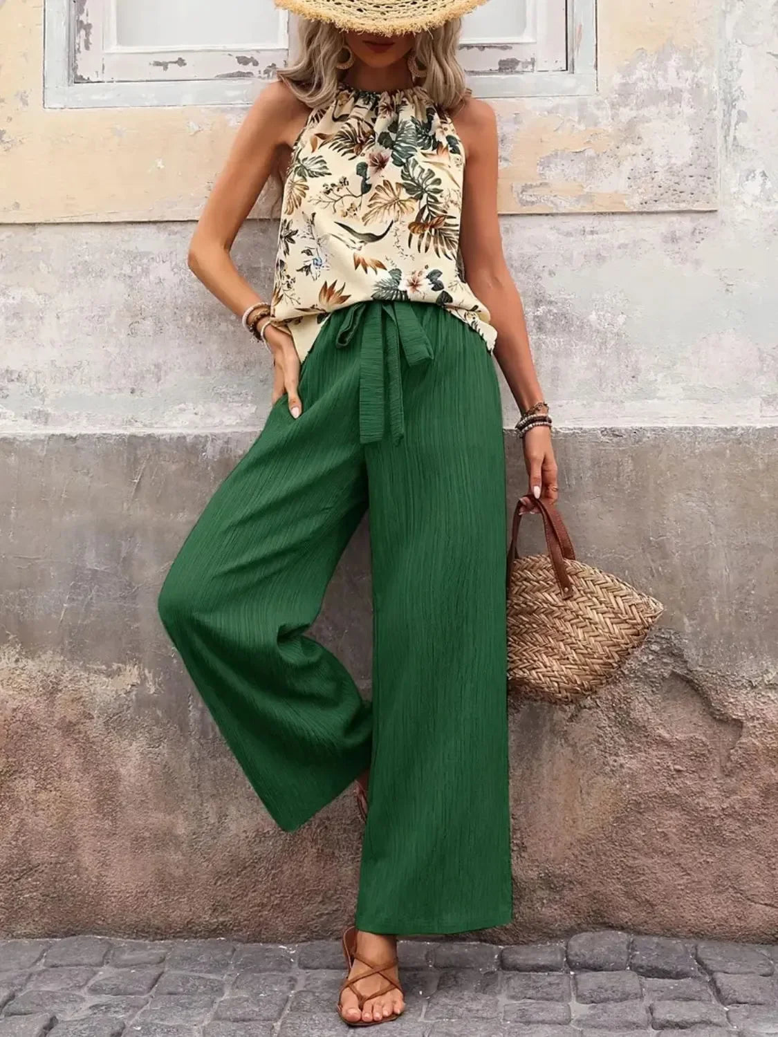 Woman wearing floral halter top, wide-leg green pants, straw hat, and sandals outdoors