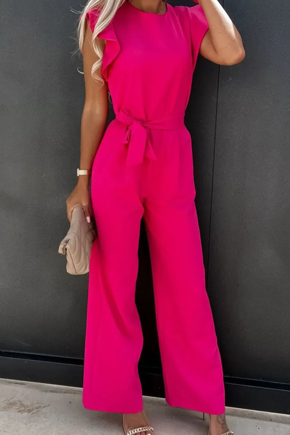 Woman wearing vibrant pink wide-leg jumpsuit with ruffled shoulders and waist tie, holding beige clutch
