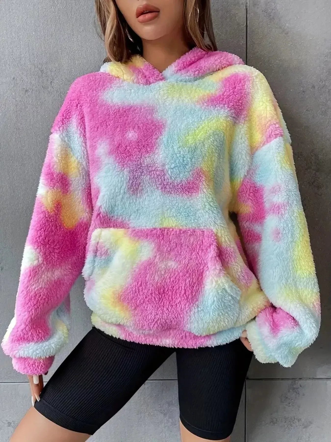 Woman wearing colorful pastel tie-dye fuzzy hoodie with black biker shorts against gray wall