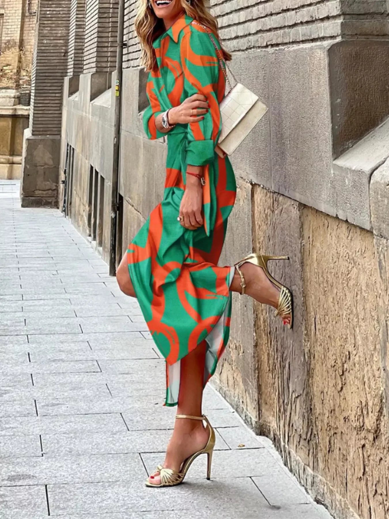 Woman in green and orange patterned midi dress and gold high heel sandals posing outdoors on sidewalk