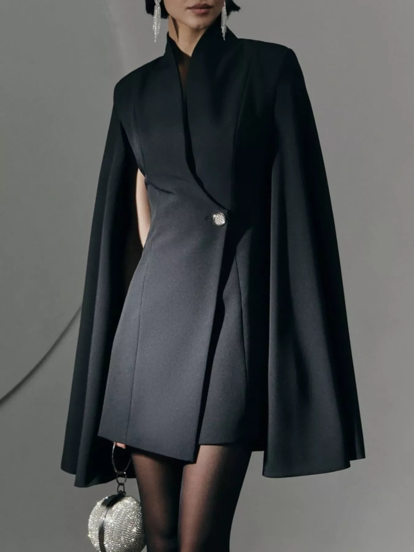Woman in black cape blazer dress with silver button, crystal earrings, and beaded clutch bag