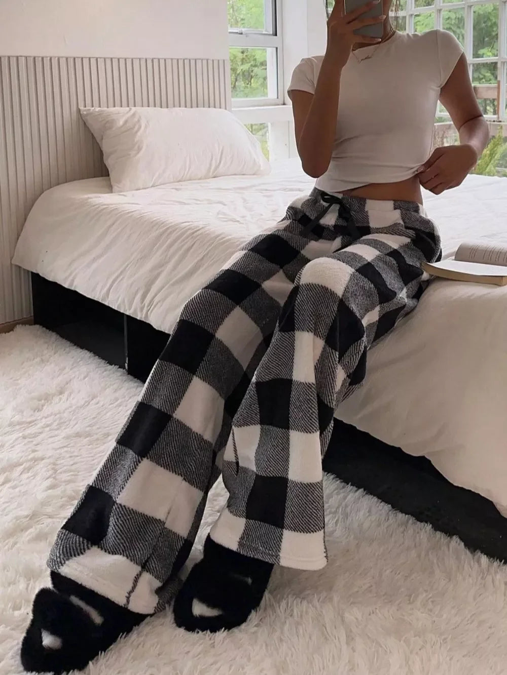 Woman wearing black and white plaid cozy lounge pants and white fitted t-shirt sitting on a bed in a bright bedroom