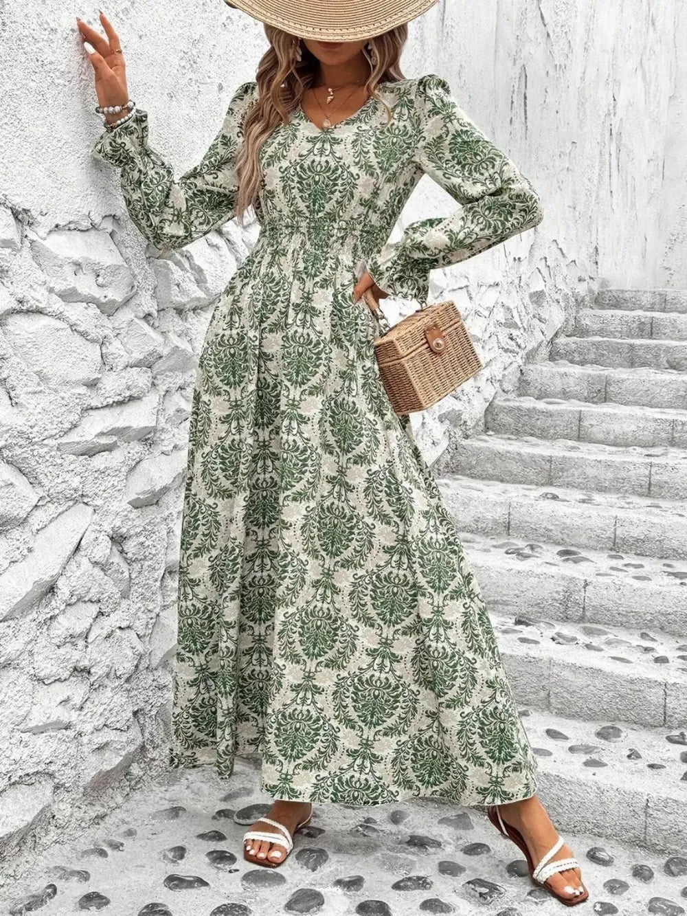 Woman in green and beige floral long-sleeve maxi dress with straw hat and woven handbag on cobblestone steps