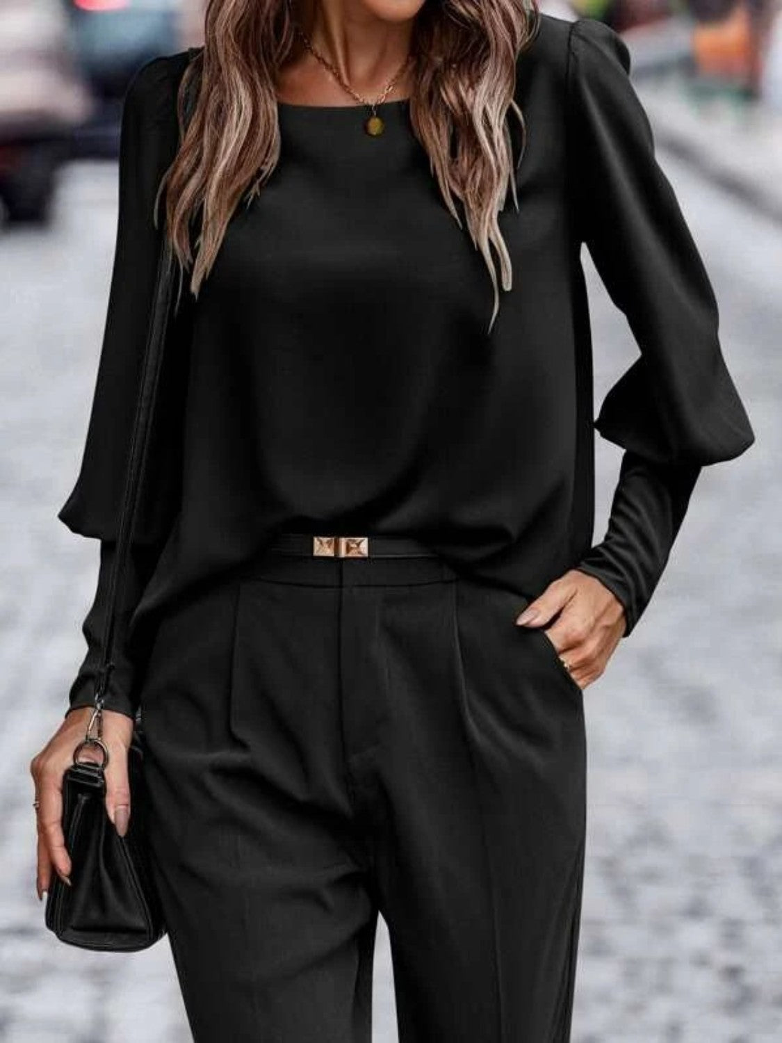 Woman wearing black long sleeve blouse and pleated pants, holding a black purse on street