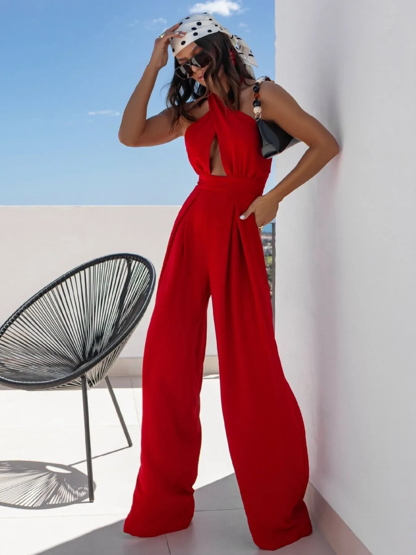 Woman in stylish red wide-leg jumpsuit with cutout, polka dot headscarf, sunglasses by white wall and black chair
