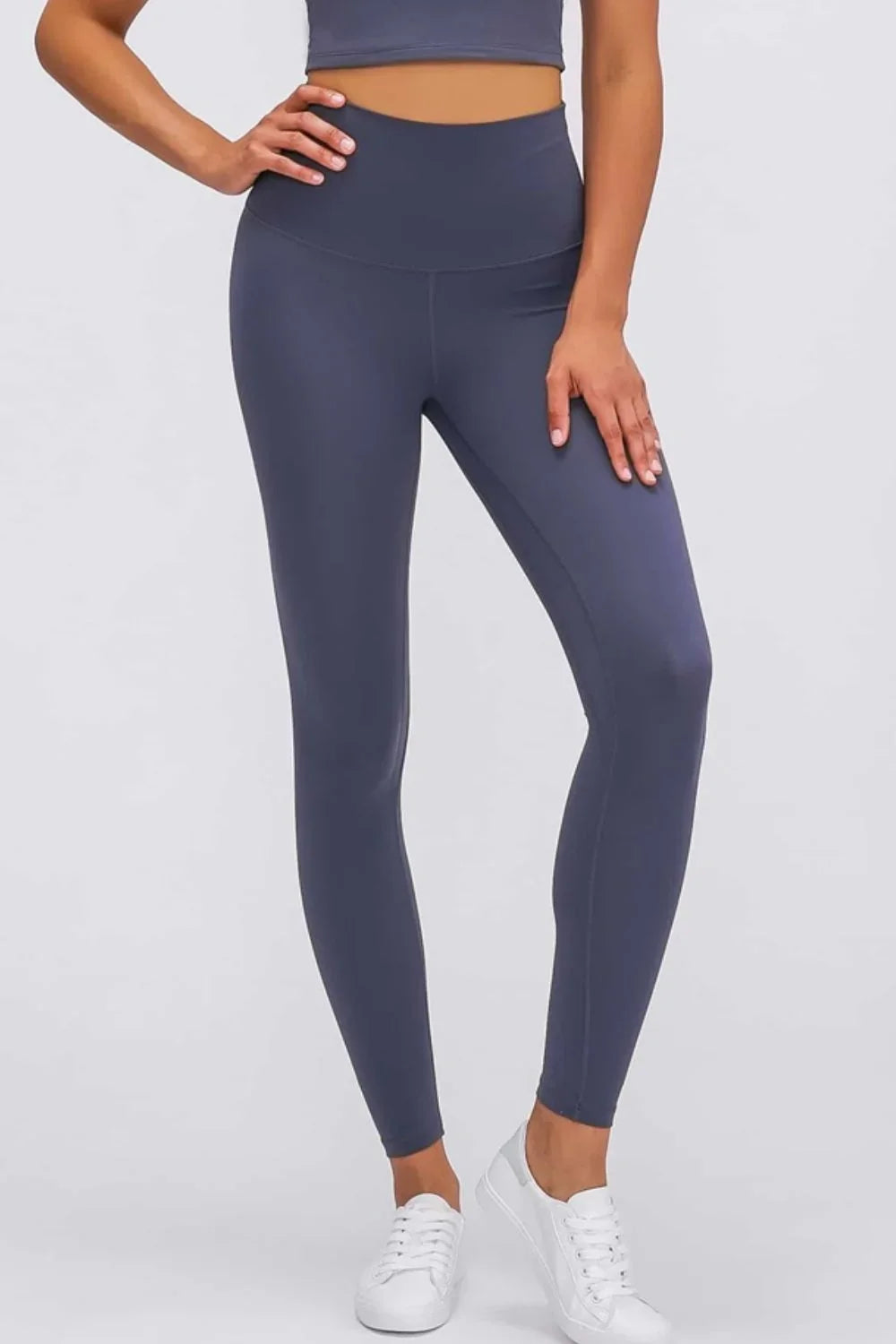 Woman wearing high-waisted dark gray athletic leggings and white sneakers on plain background