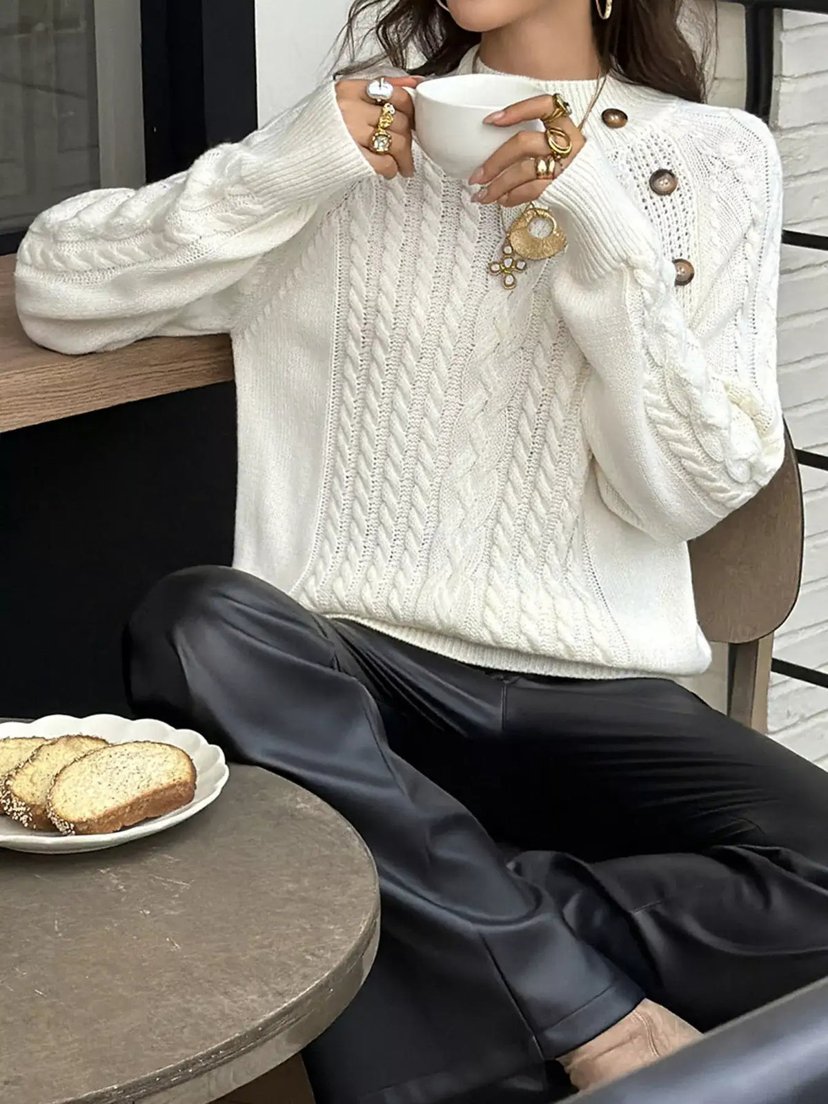 Woman wearing an Evie's Dreamland white mock neck cable-knit sweater with shoulder buttons and black leather pants.