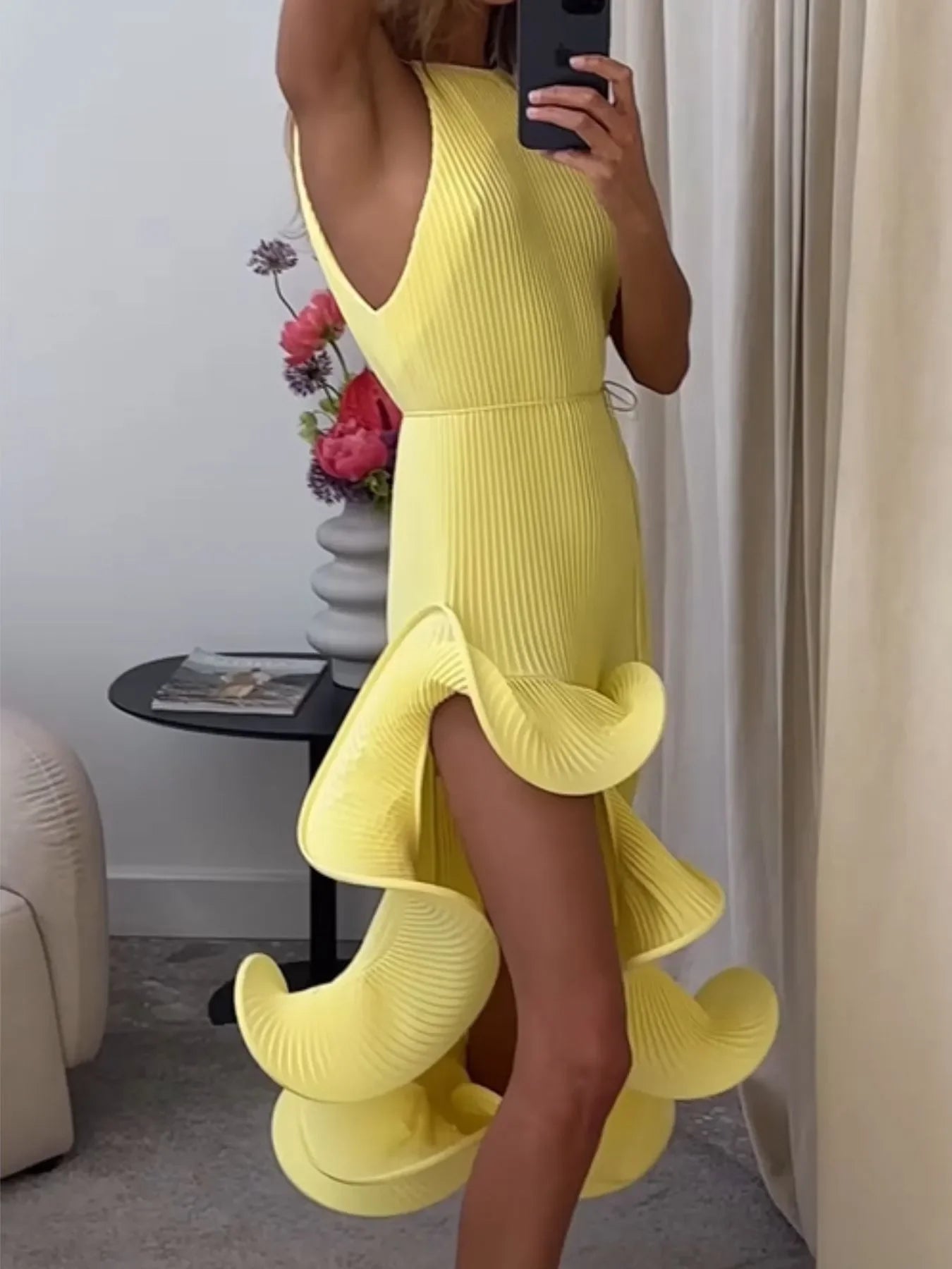 Woman wearing sleeveless yellow ruffle dress in modern room with flowers and magazine table