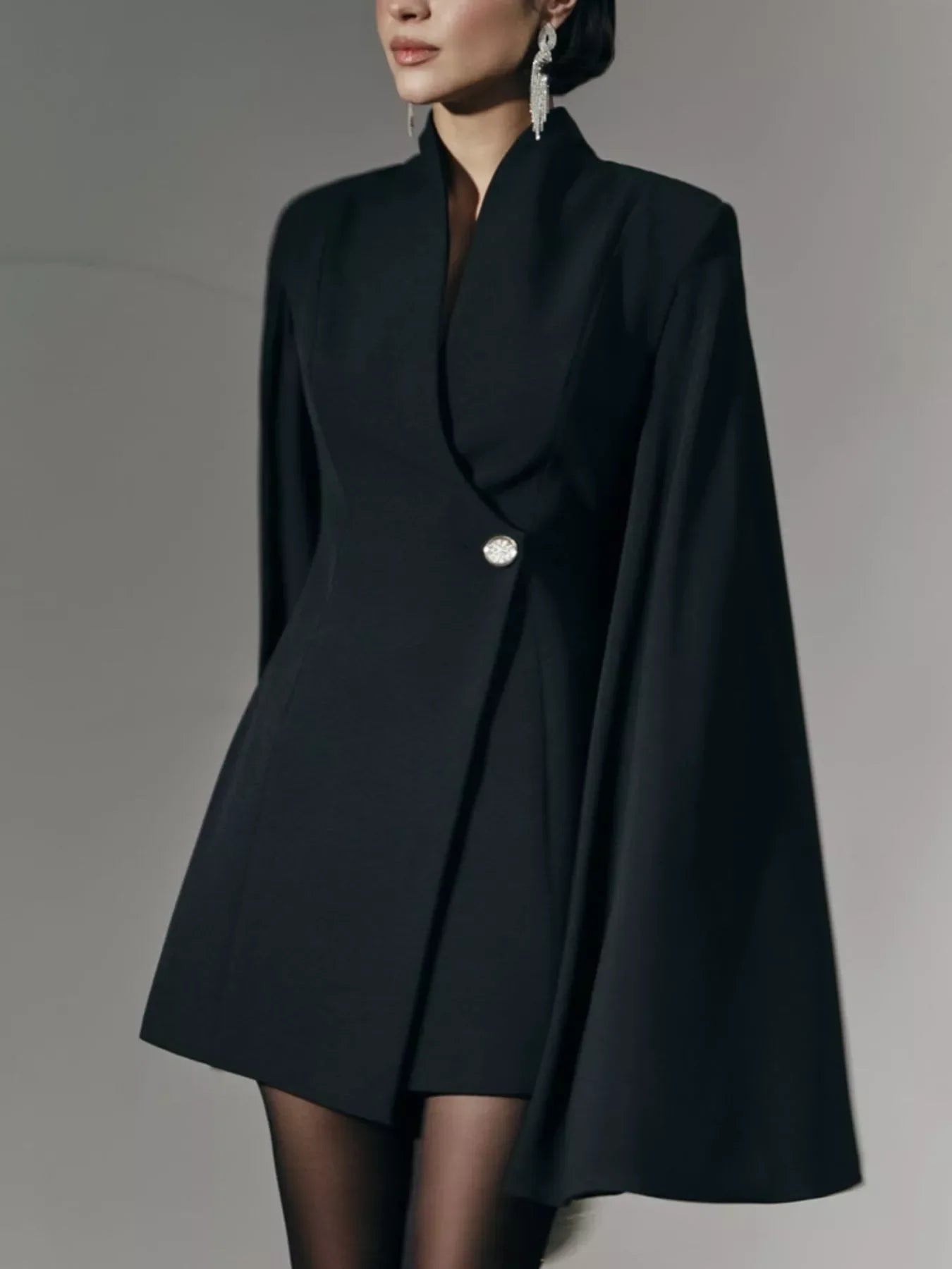 Woman wearing chic black cape blazer dress with single decorative button and statement earrings