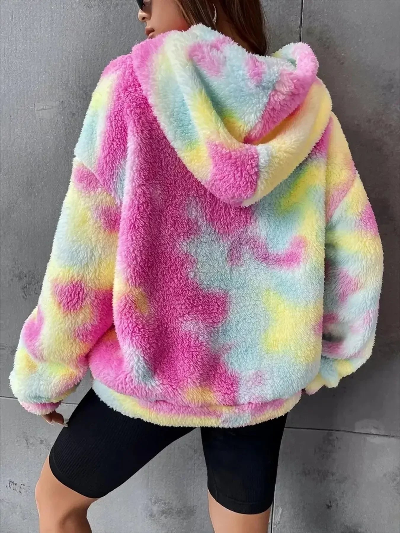 Woman wearing colorful pastel tie-dye fluffy hoodie with hood and black biker shorts against gray wall