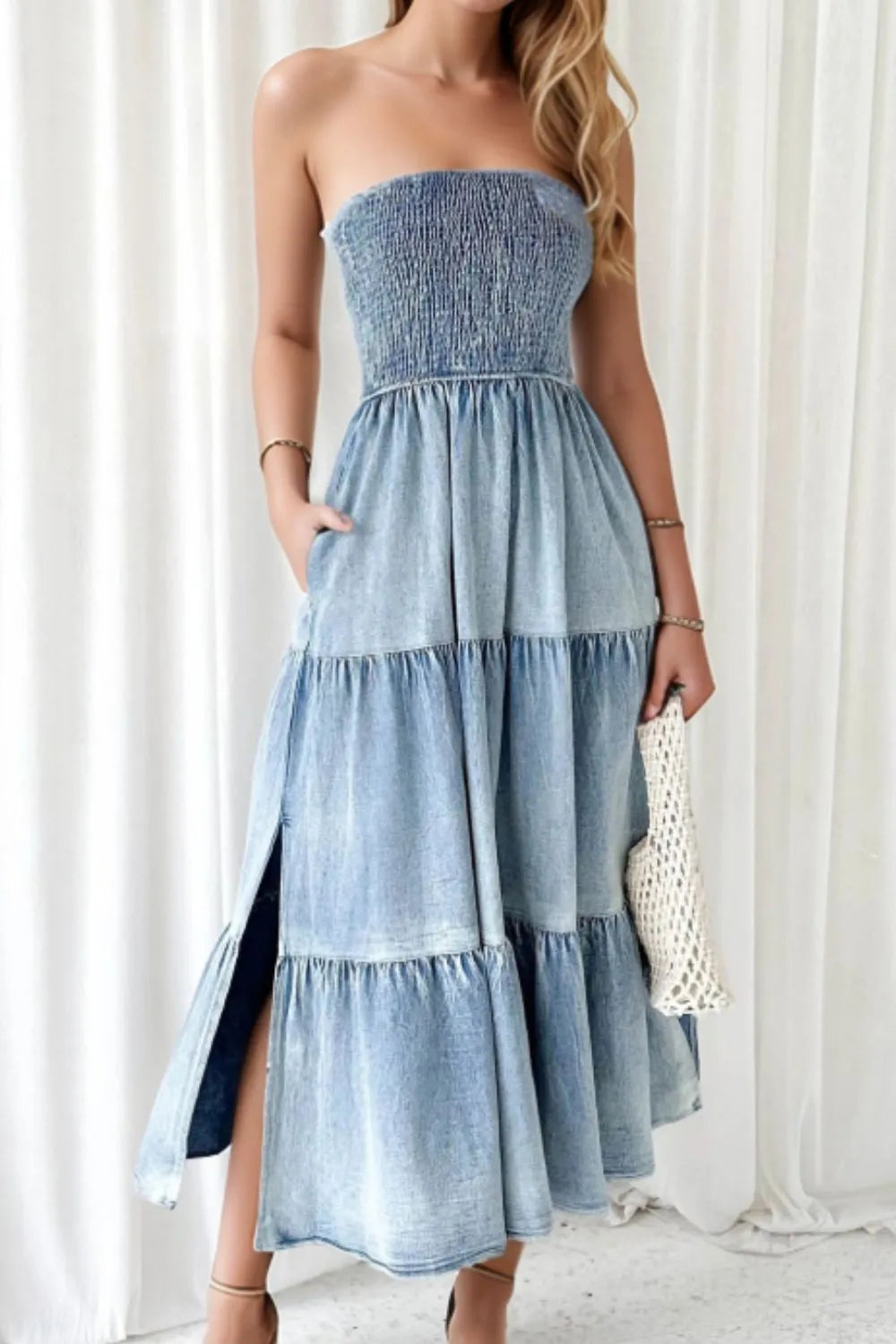 Woman wearing a strapless denim midi dress with tiered skirt and side slit, holding a woven clutch
