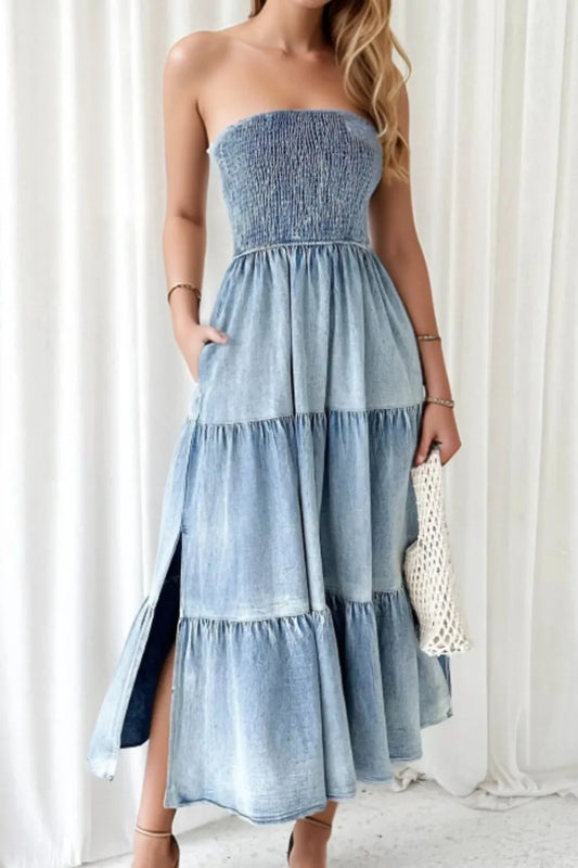Woman wearing a strapless denim midi dress with tiered skirt and side slit, holding a woven clutch