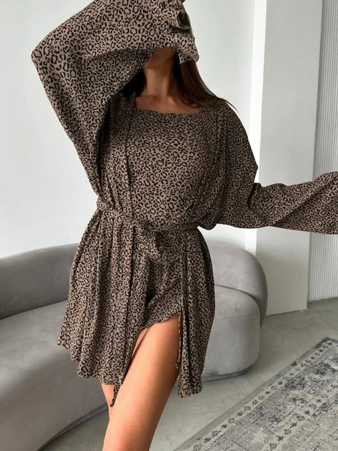Woman wearing a leopard print long-sleeve dress with a slit, posing indoors by a gray couch