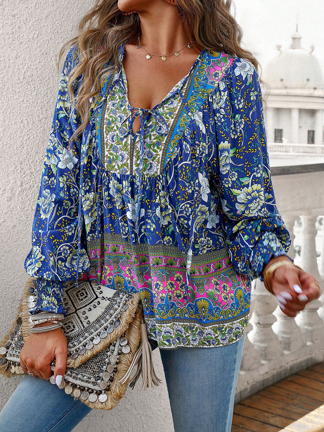 Boho floral blouse with long sleeves, V-neck tie, and vibrant print, paired with jeans and fringe clutch