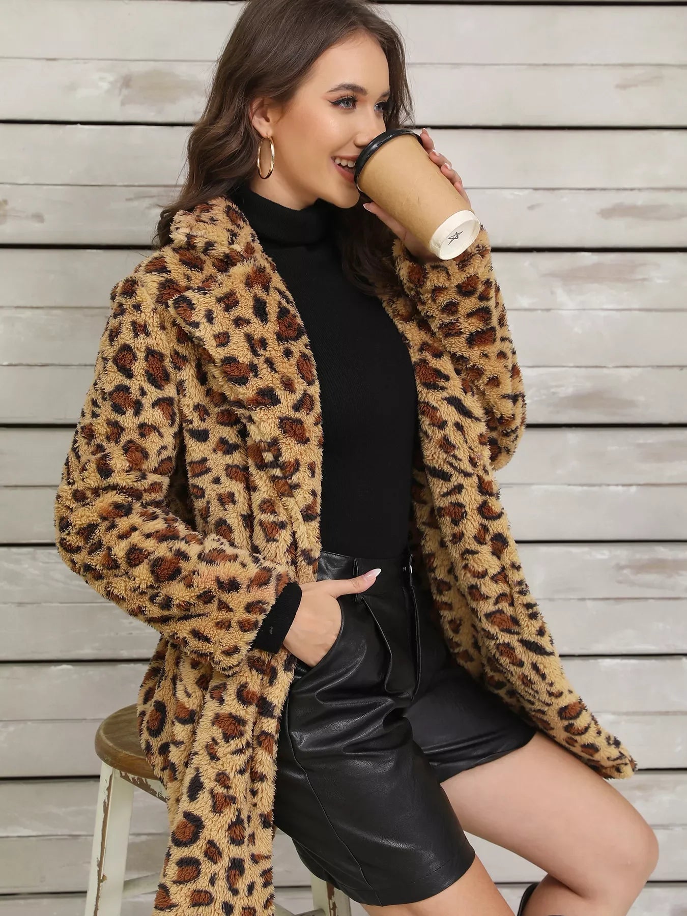 Woman wearing leopard print faux fur coat, black turtleneck, and leather shorts sipping coffee