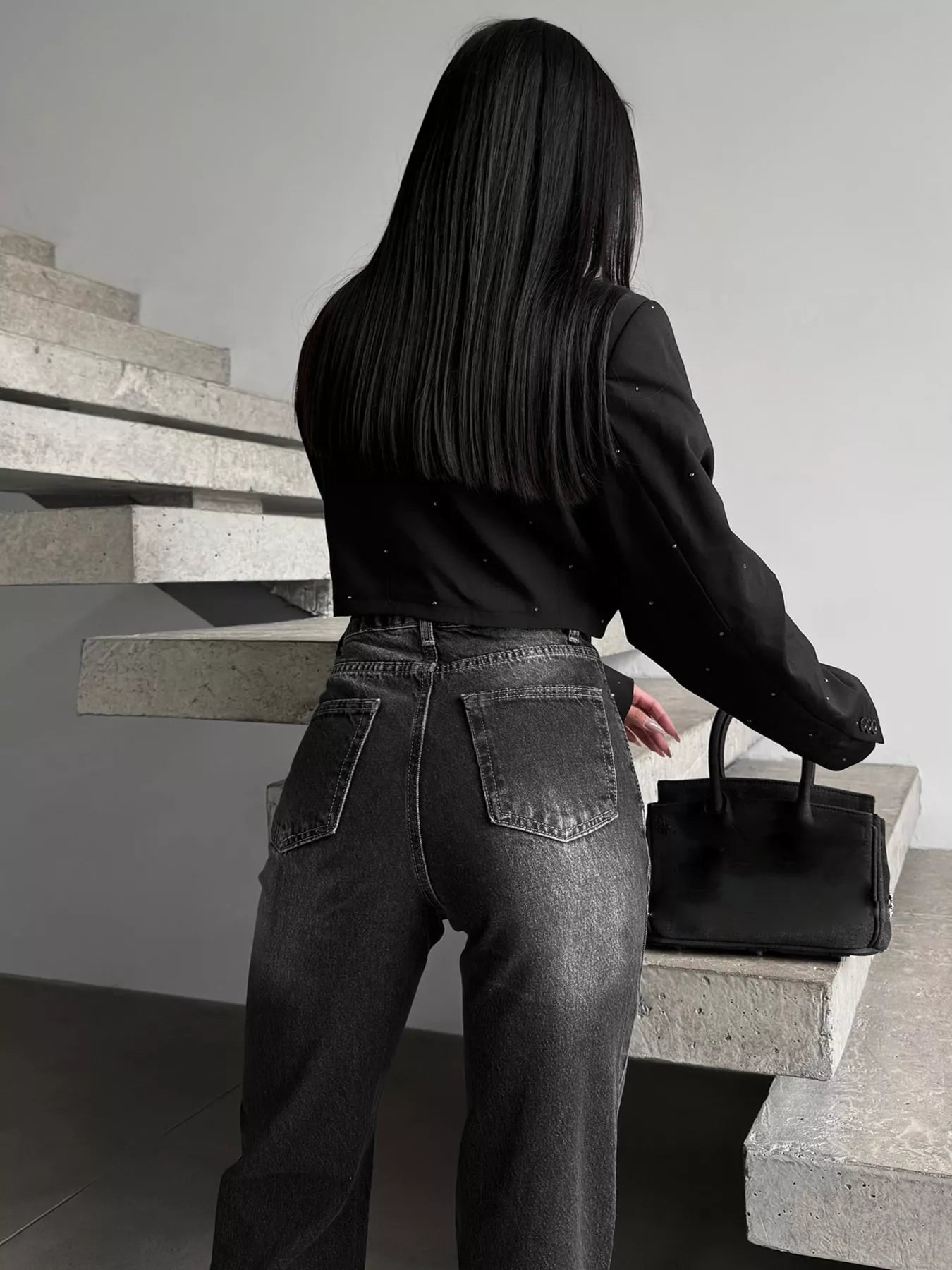 Woman in black cropped blazer and high-waisted faded jeans holding a chic black handbag