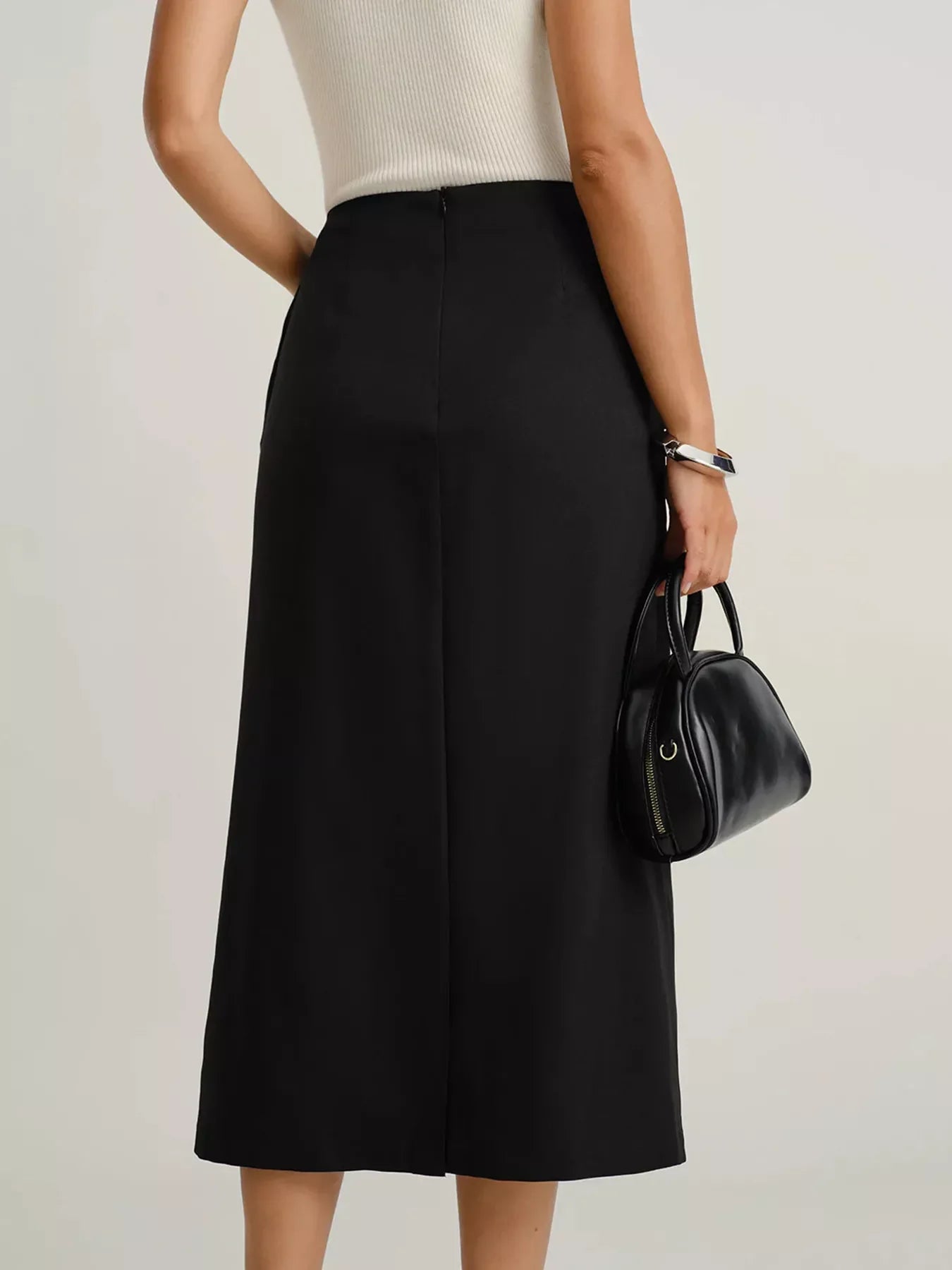 Back view of woman wearing black midi skirt and white sleeveless top holding black leather handbag