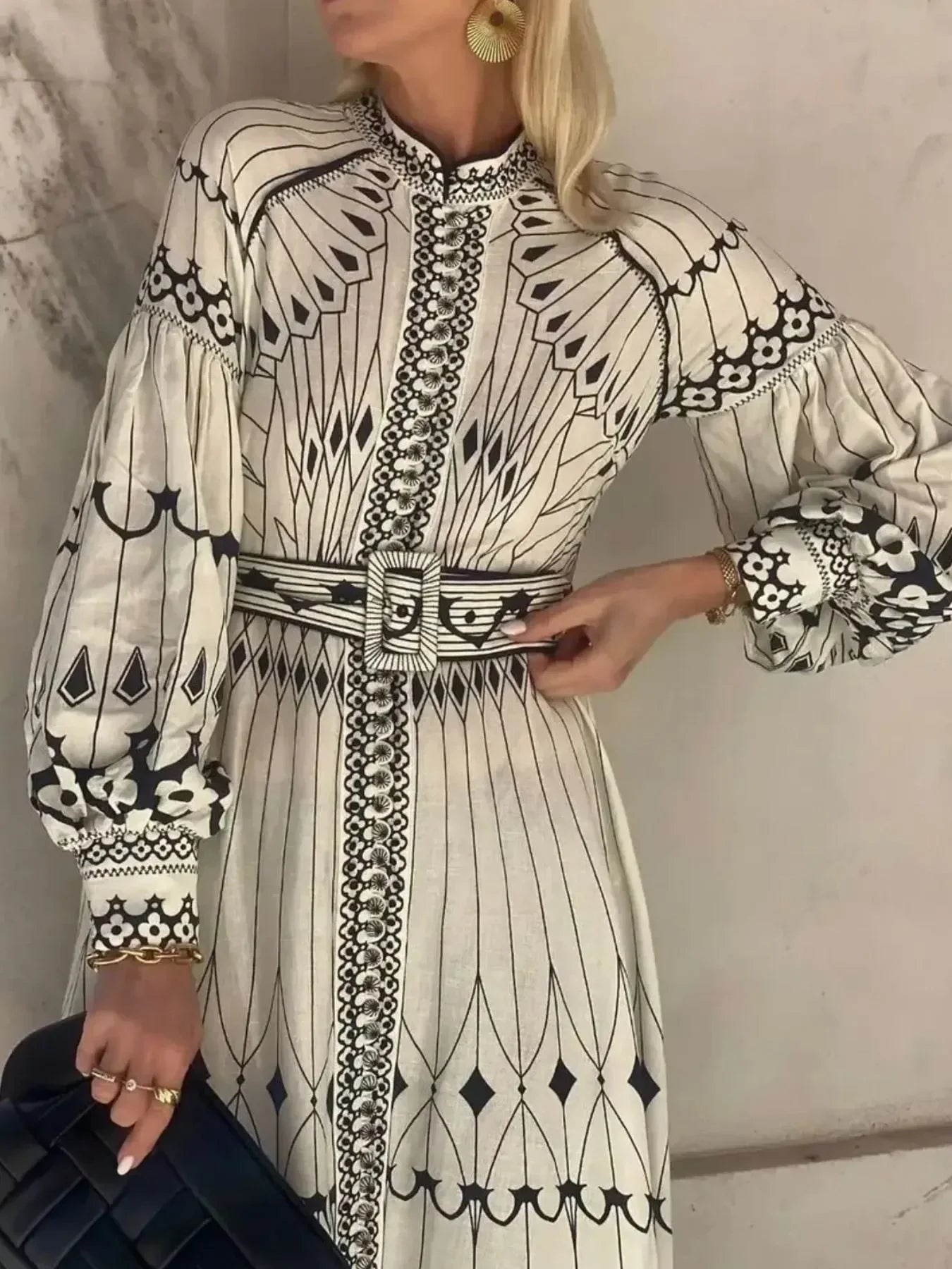 Woman wearing elegant cream dress with black geometric and floral patterns, puffed sleeves, belt, gold jewelry, and holding black quilted handbag