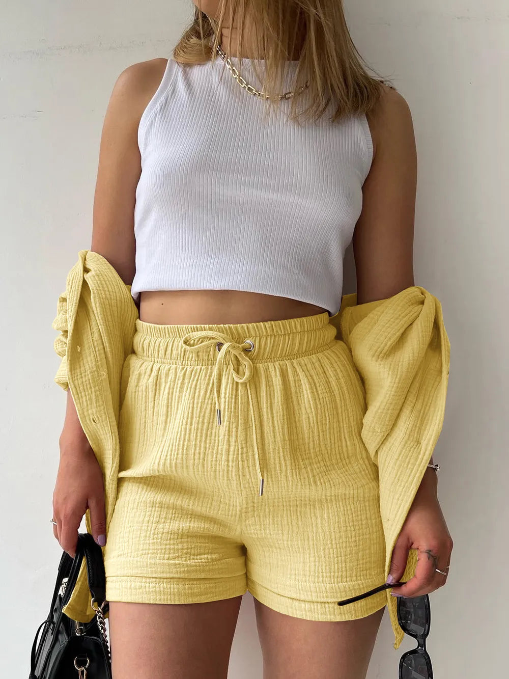 Woman wearing a white ribbed crop top and matching yellow textured shorts set, holding accessories.