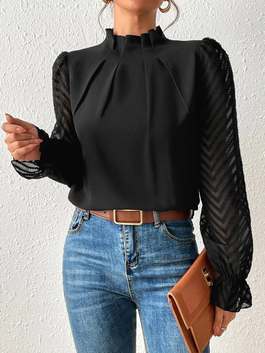 Woman wearing a chic black blouse with sheer long sleeves, blue jeans, and holding a tan clutch.