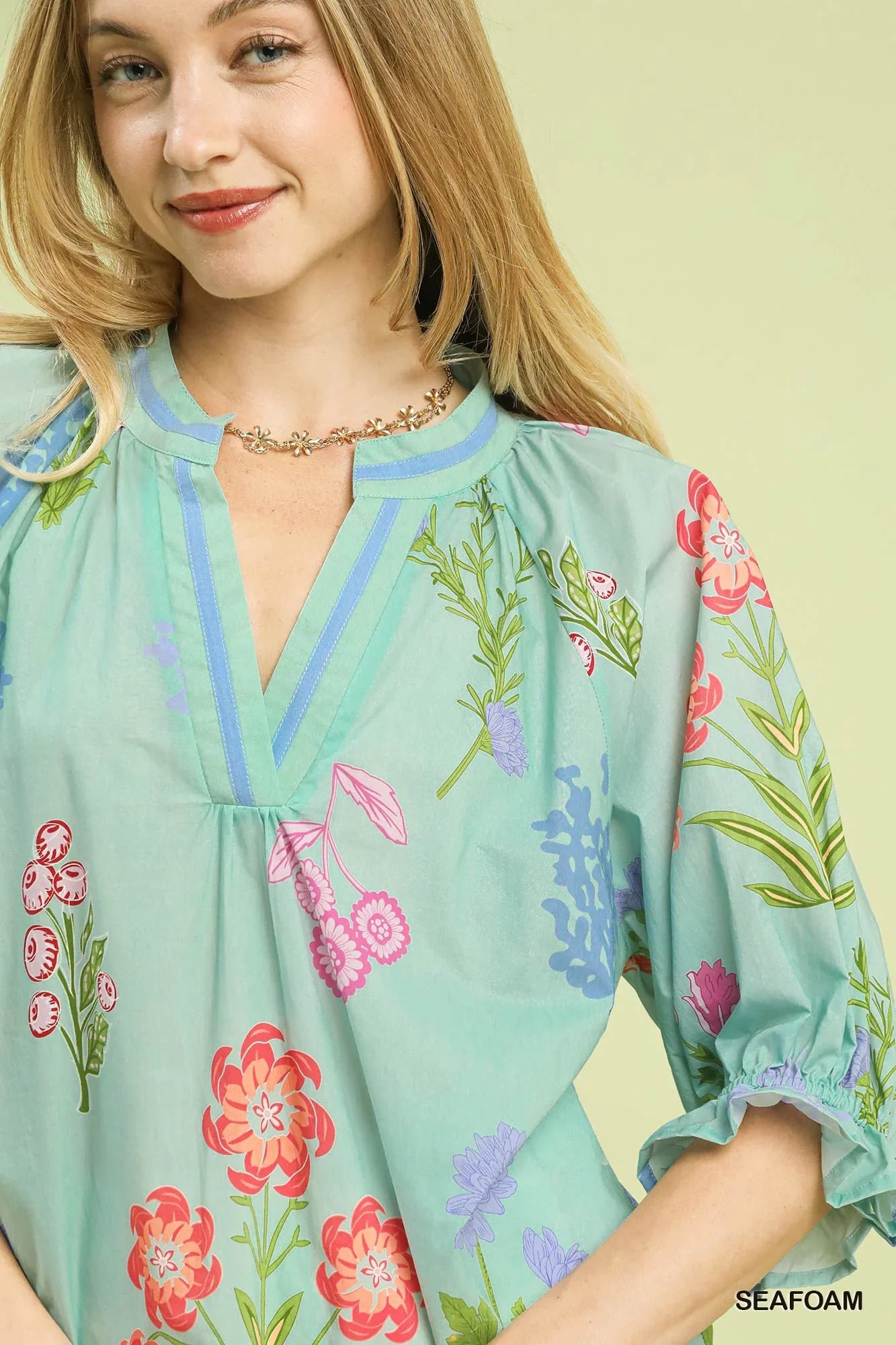 Woman wearing a seafoam green floral blouse with ruffled sleeves and a gold flower necklace