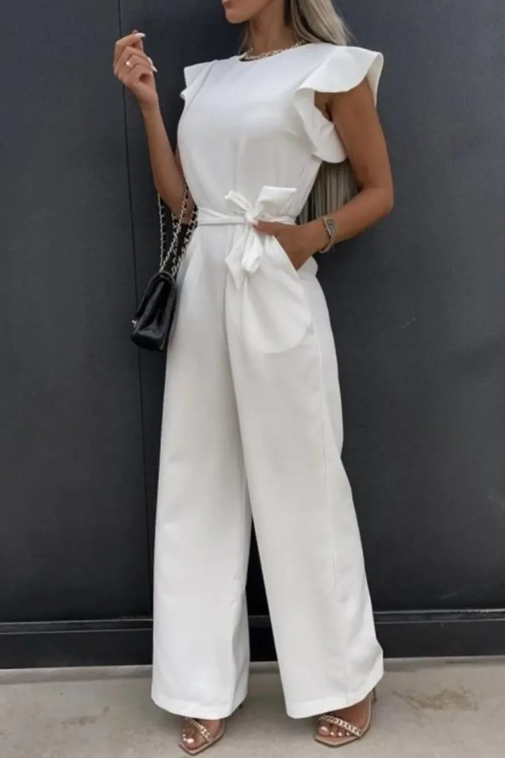 Woman wearing white ruffle sleeve jumpsuit with wide-leg pants and tied waist belt, carrying black purse