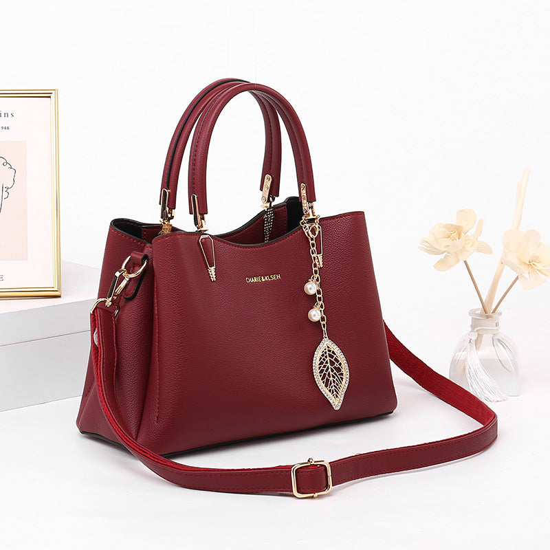 Stylish burgundy leather handbag with top handles, gold hardware, and leaf charm, on white desk