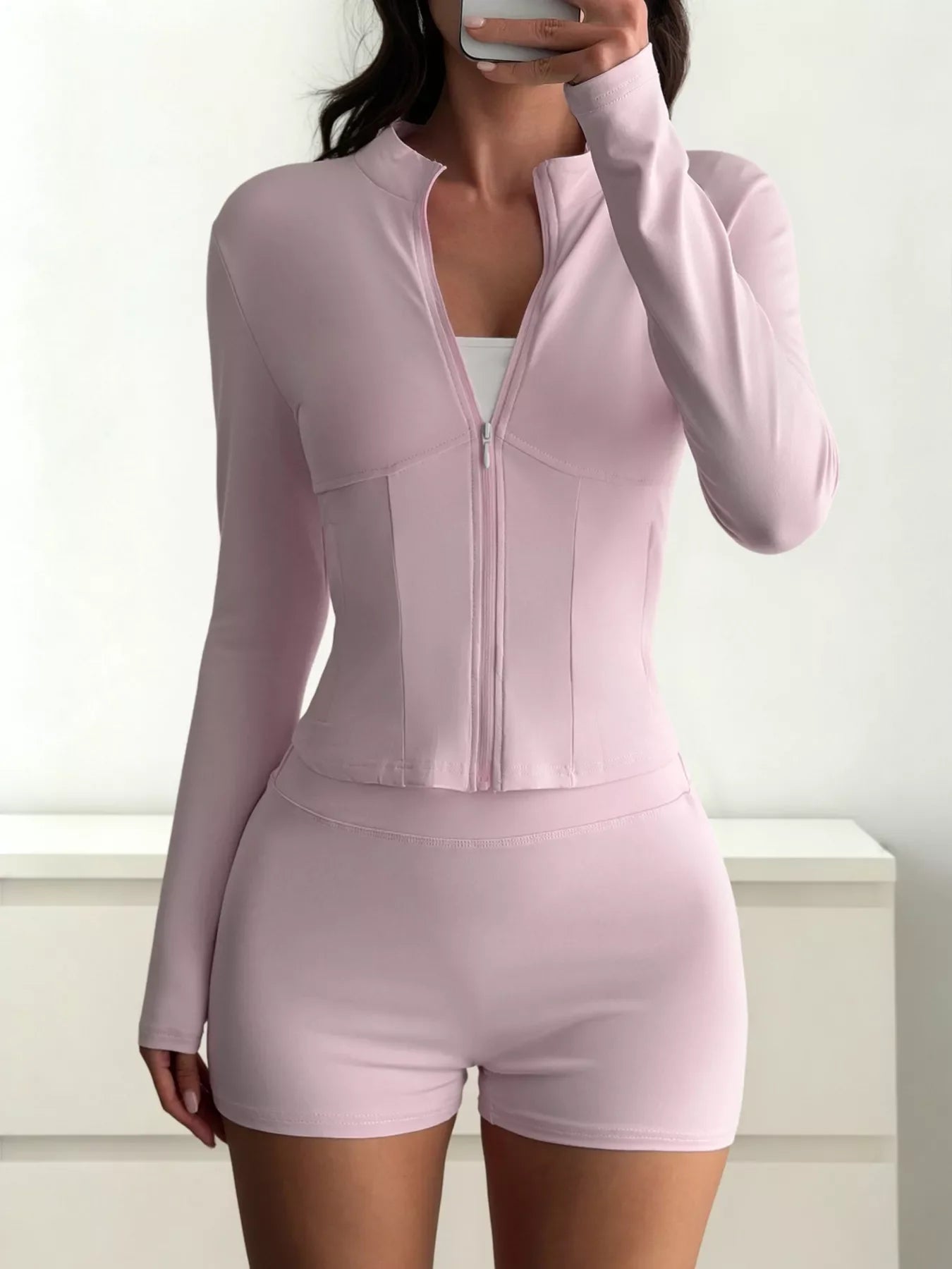 Woman wearing light pink zip-up long sleeve top and matching shorts in minimal indoor setting