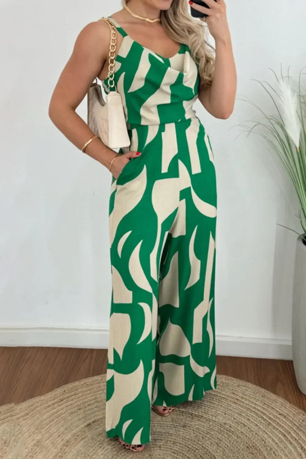 Woman wearing green and beige abstract print sleeveless jumpsuit with wide legs, holding beige purse
