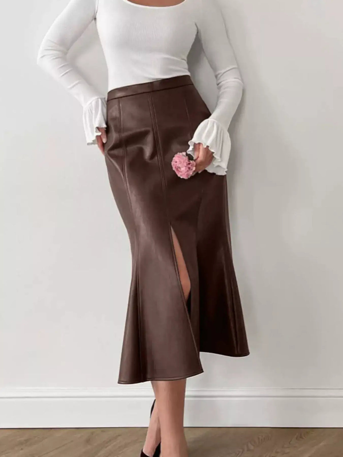 Woman wearing a brown faux leather mermaid midi skirt with a front slit and a white long-sleeve top.
