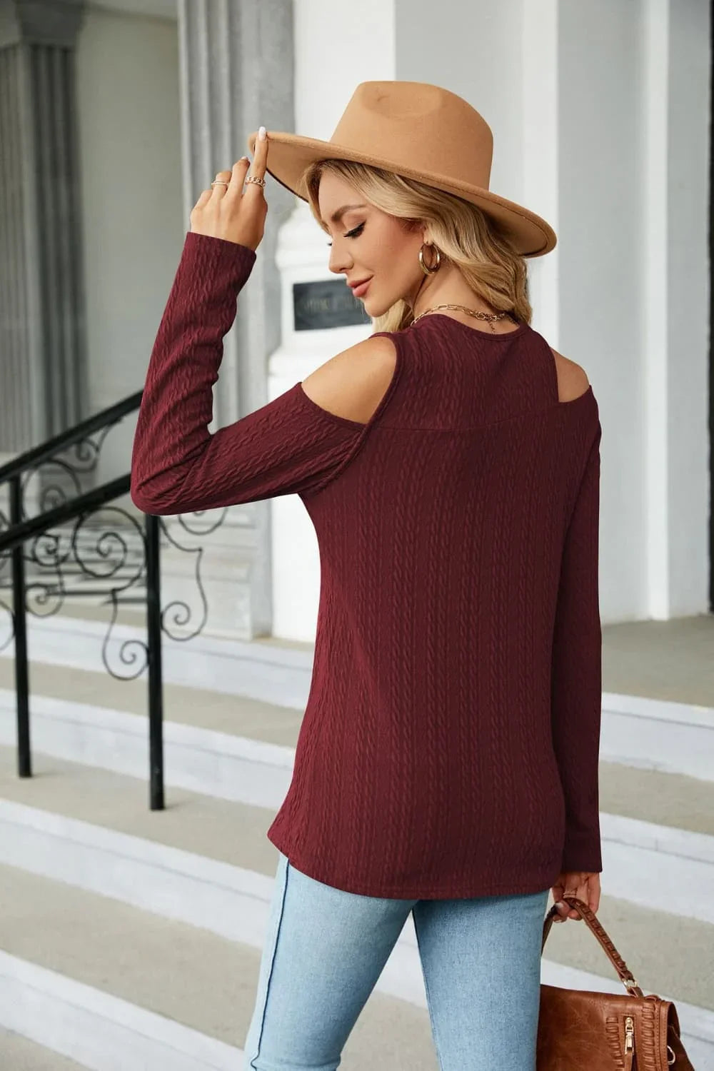 Woman in a maroon cold shoulder sweater, light jeans, tan hat, holding brown handbag outdoors