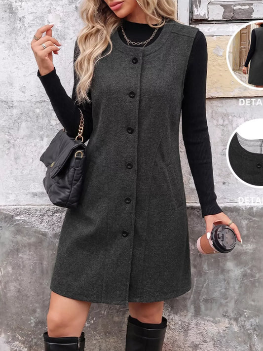 Woman wearing a dark grey button-down sleeveless dress over a black long-sleeve top.