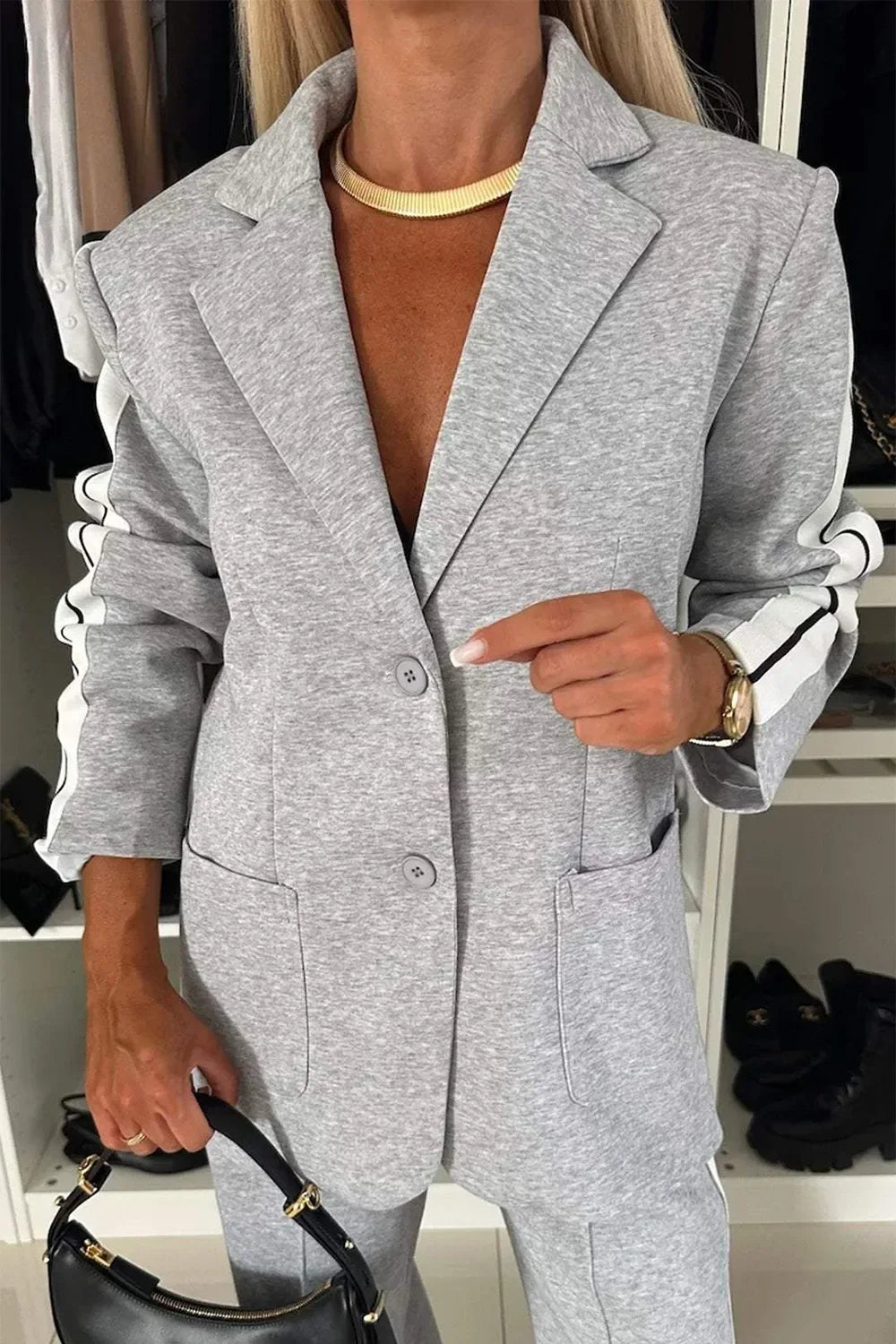 Woman wearing light gray casual blazer with white striped sleeves, holding black handbag, gold necklace, and watch