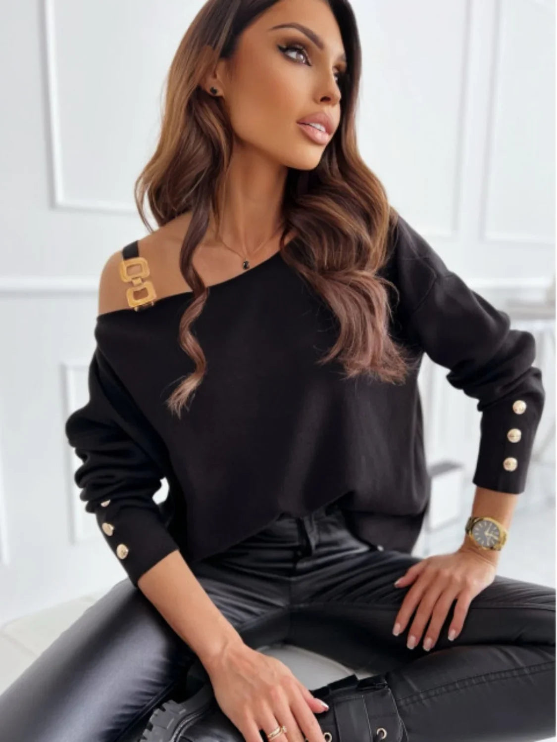 Woman in chic black off-shoulder sweater with gold buttons and faux leather pants sitting stylishly