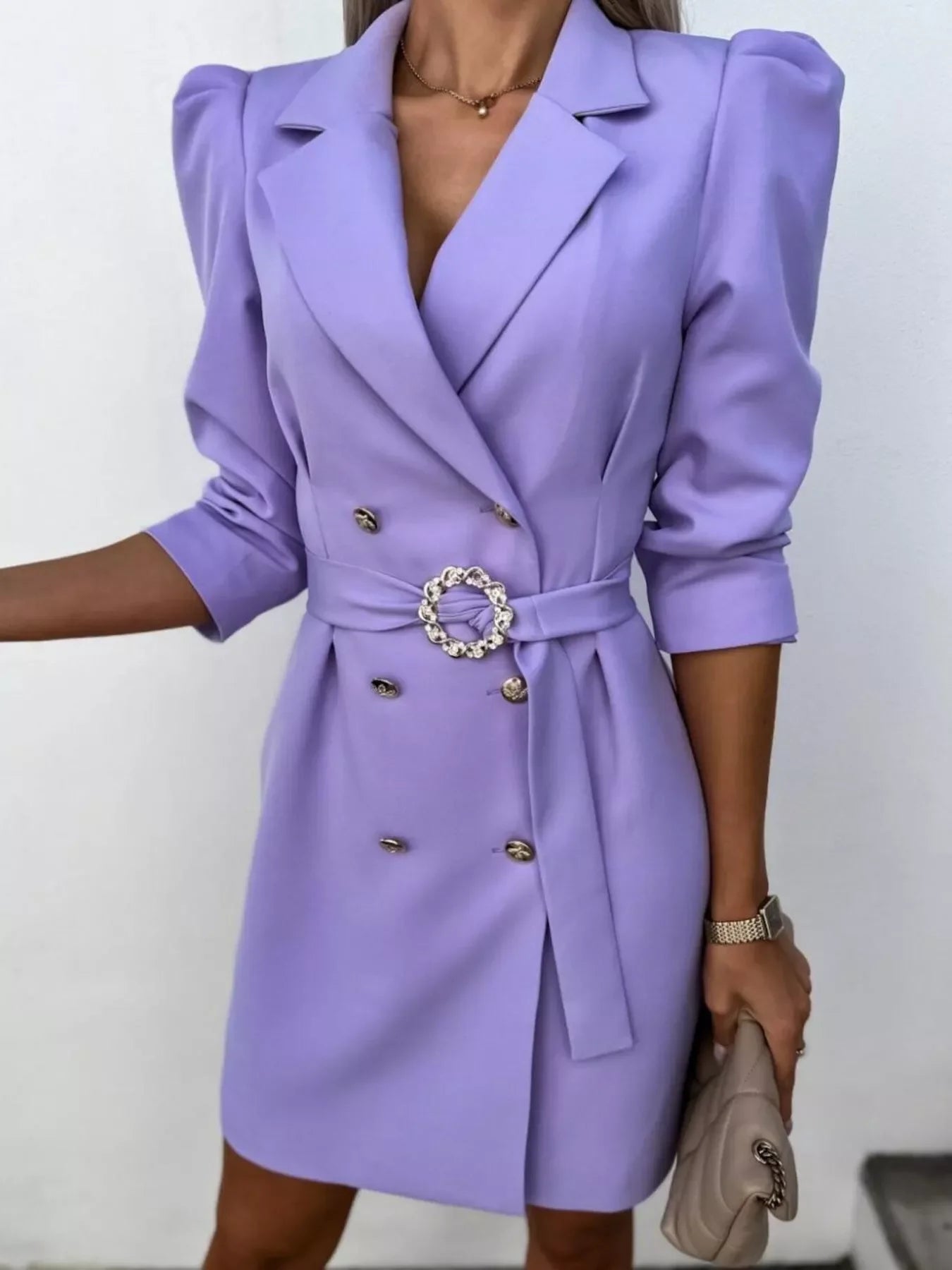 Woman wearing a lavender double-breasted blazer dress with puffed sleeves and a decorative belt holding a beige quilted handbag