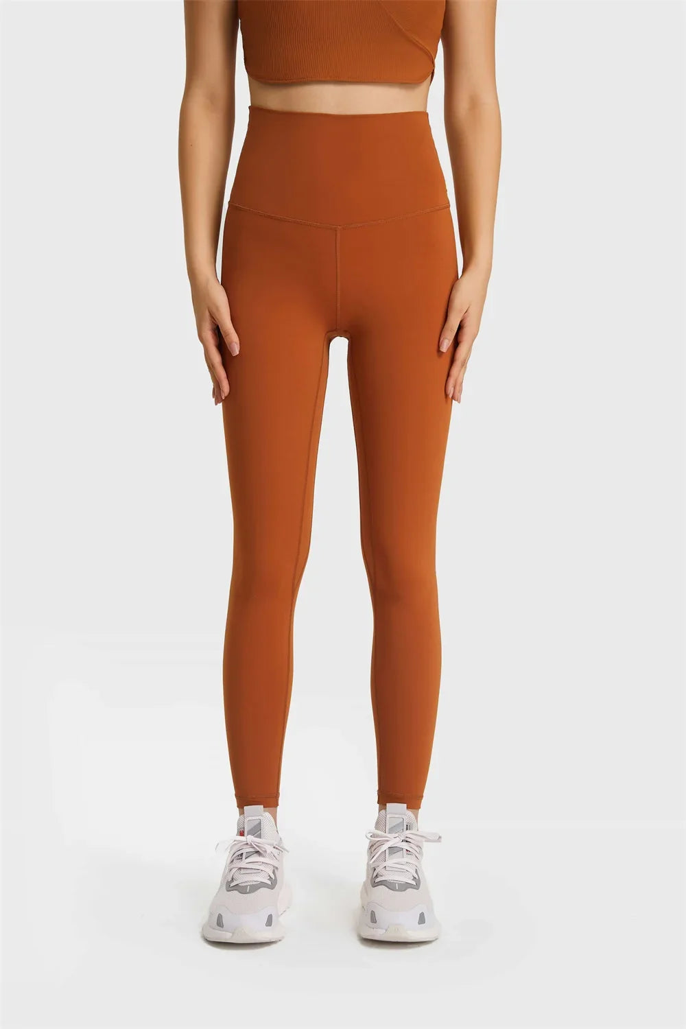 Front view of woman wearing rust-colored high-waisted leggings and white sneakers on gray background
