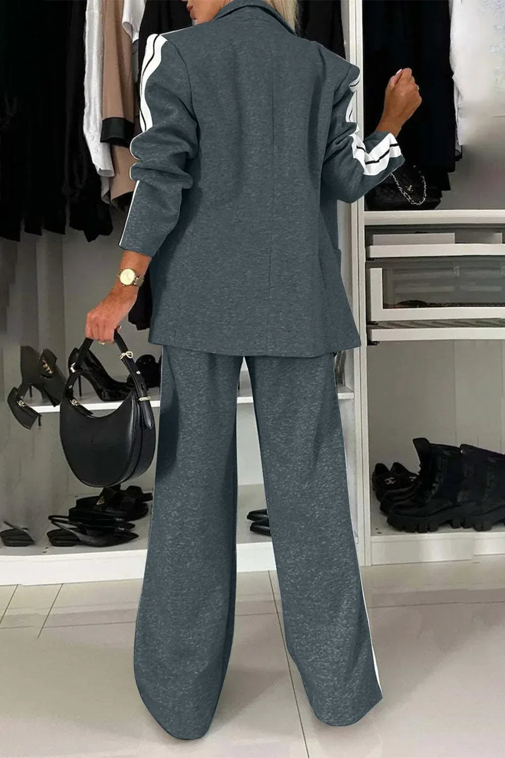 Woman wearing gray pantsuit with white stripes on sleeves and holding black handbag in closet