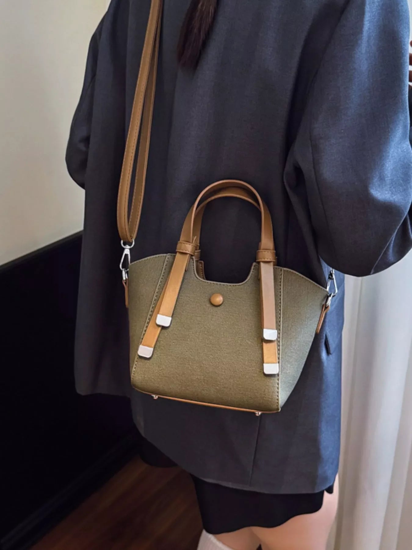 Woman wearing dark blazer holding khaki and tan leather small handbag with shoulder strap indoors