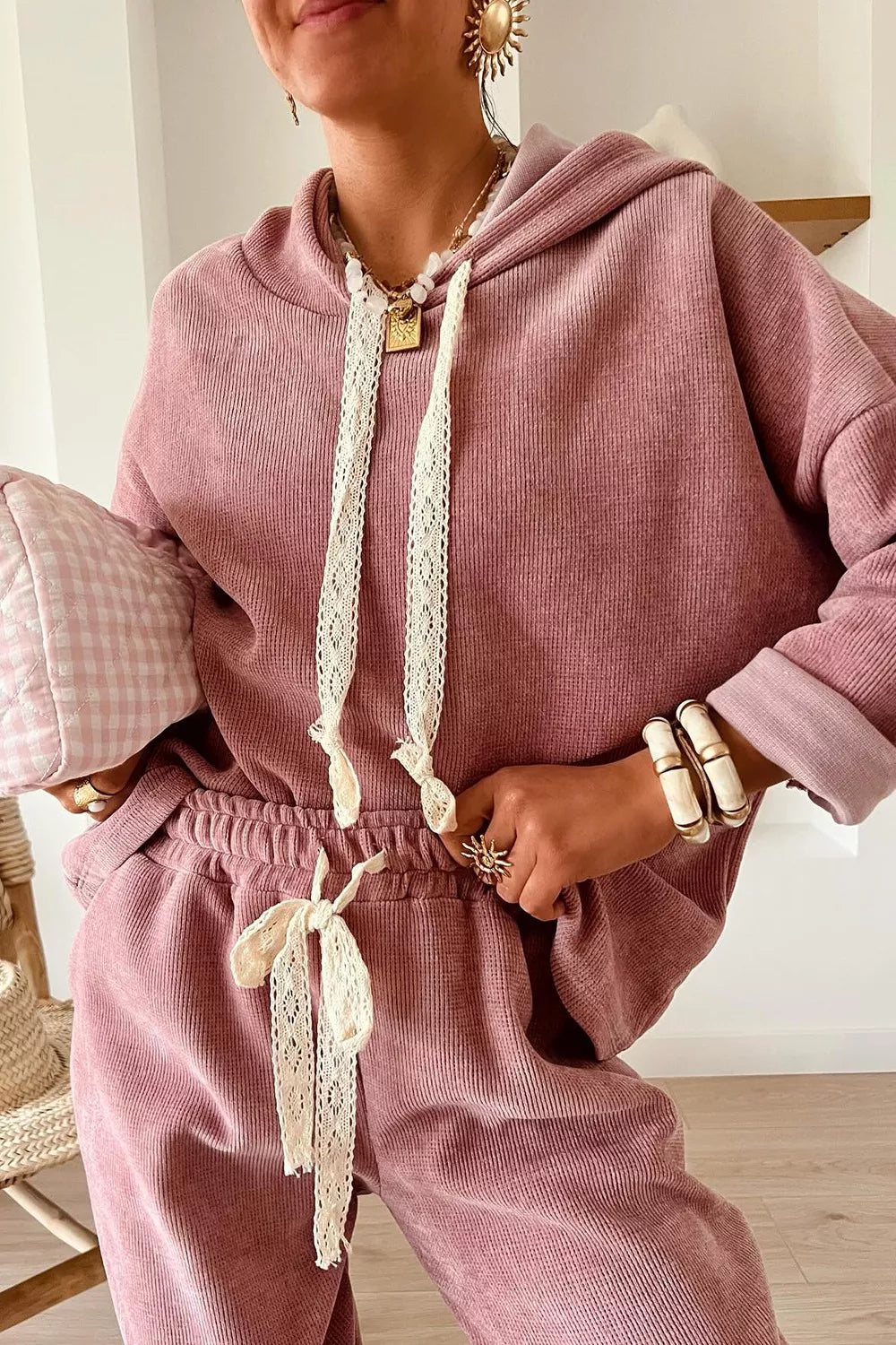 Woman wearing a cozy mauve lounge set with lace drawstrings, accessorized with chunky jewelry and a pink checkered clutch.