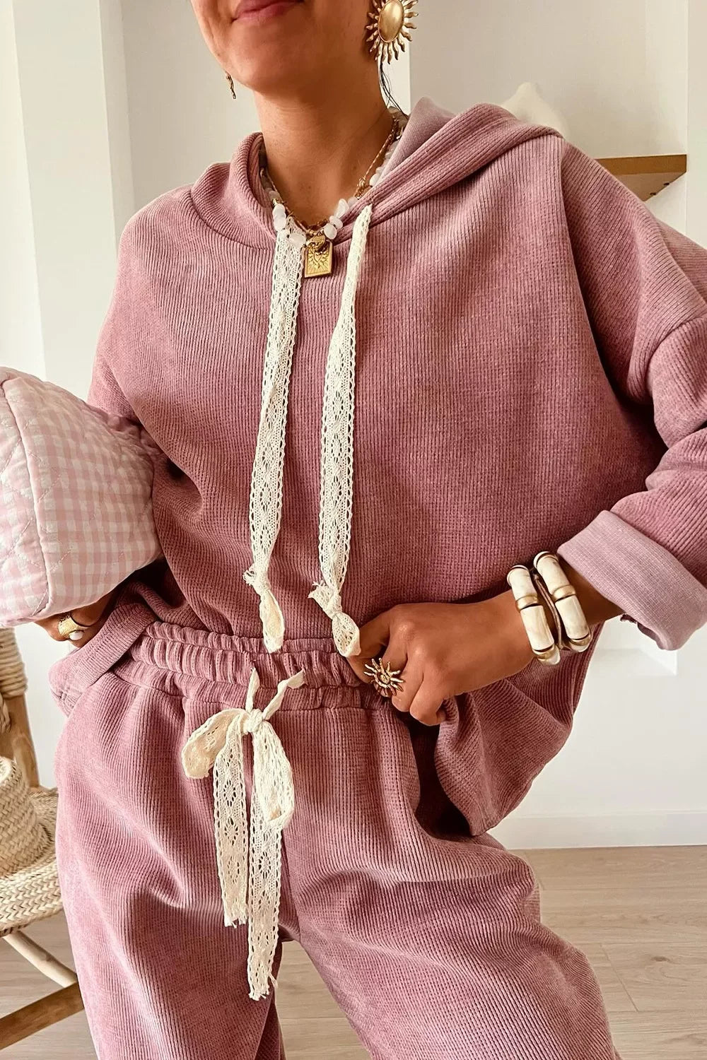 Woman wearing a cozy mauve lounge set with lace drawstrings, accessorized with chunky jewelry and a pink checkered clutch.