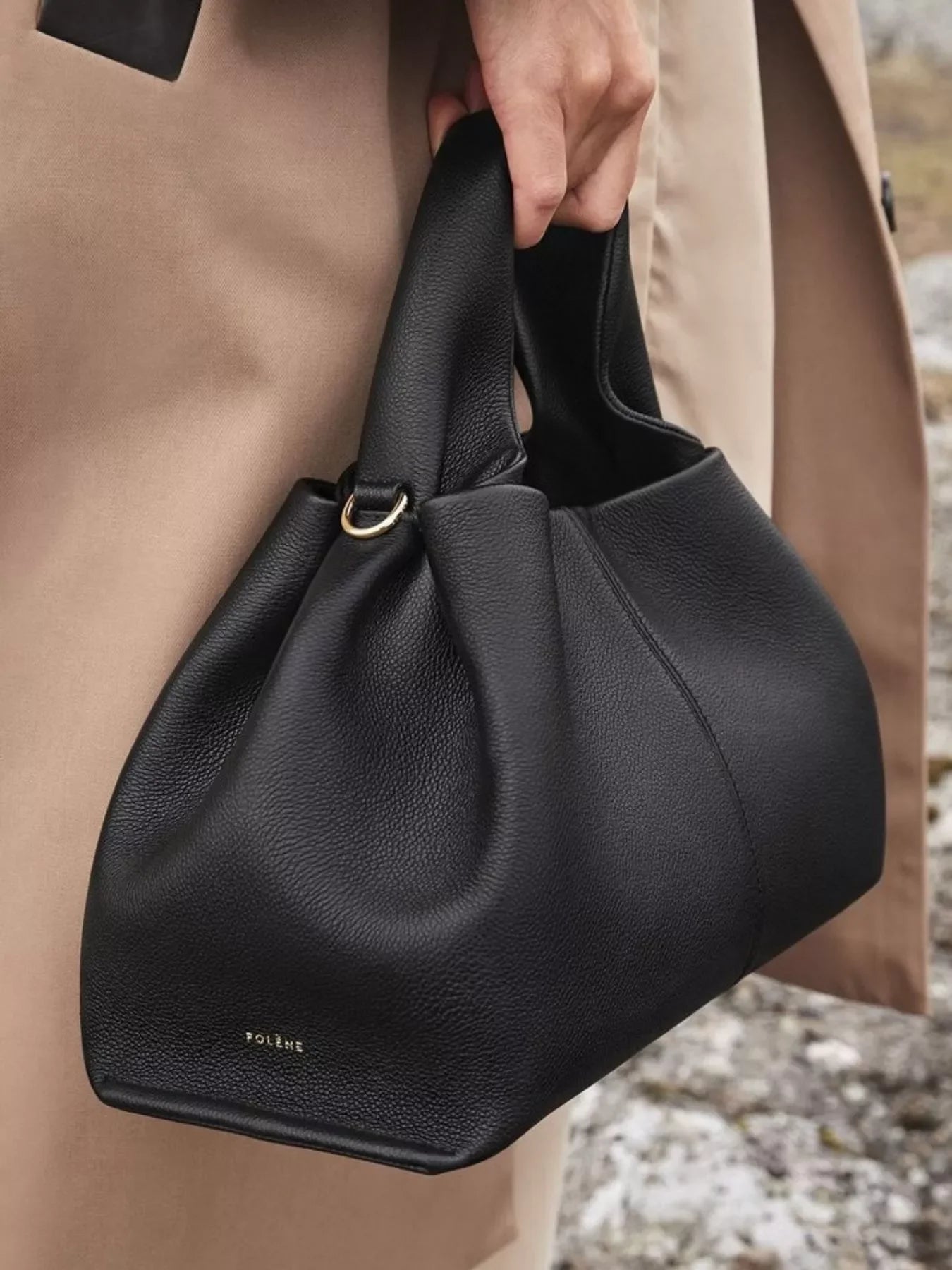 Hand holding black leather Polène handbag with gold ring detail against beige coat background
