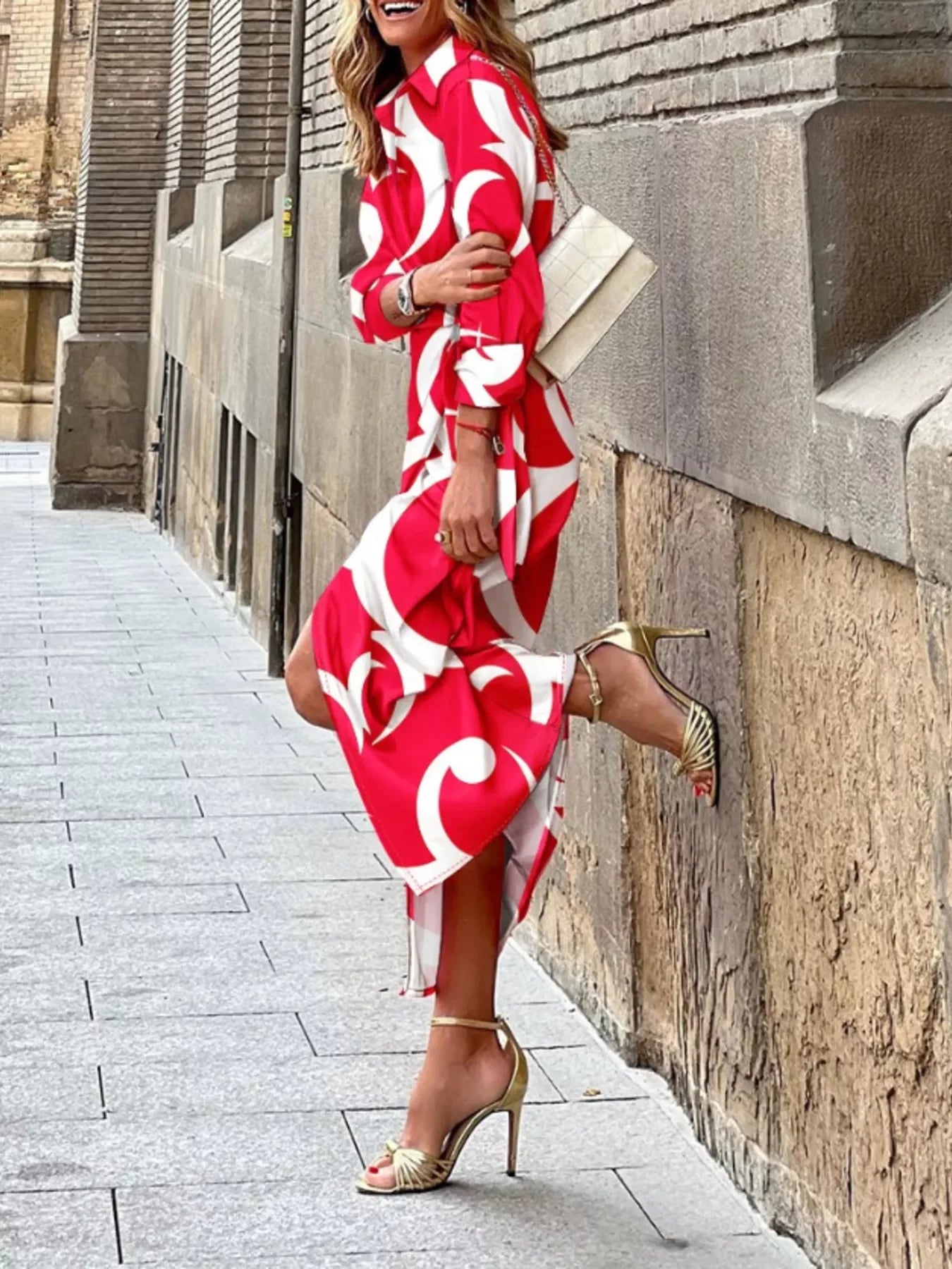 Woman in red and white patterned dress wearing gold high heel sandals and carrying beige handbag on city street