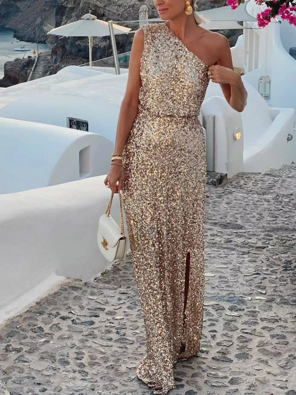 Woman wearing a gold sequin one-shoulder maxi dress with a side slit, styled with a white handbag.