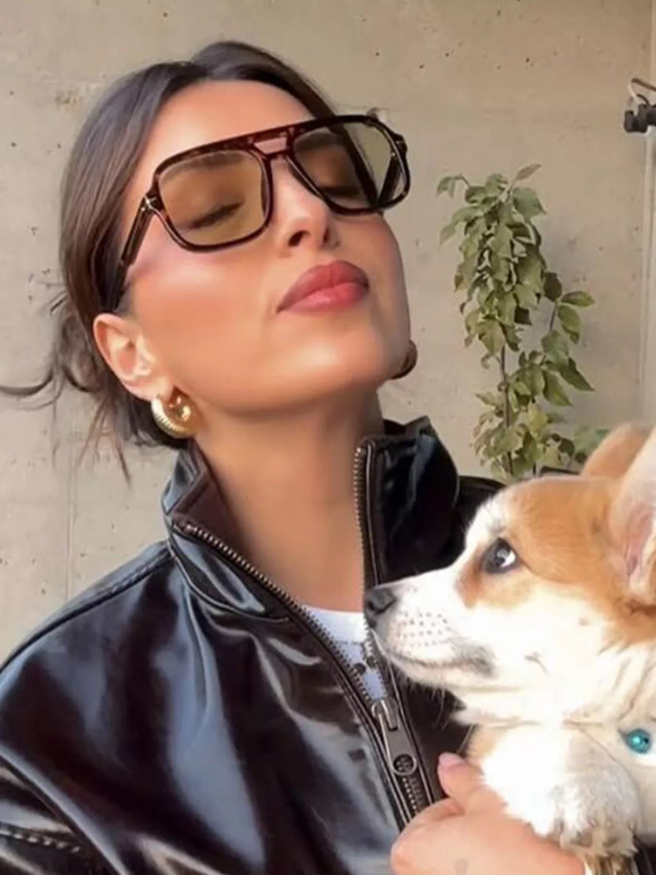 Woman wearing oversized square sunglasses, gold hoop earrings, and a black patent leather jacket.