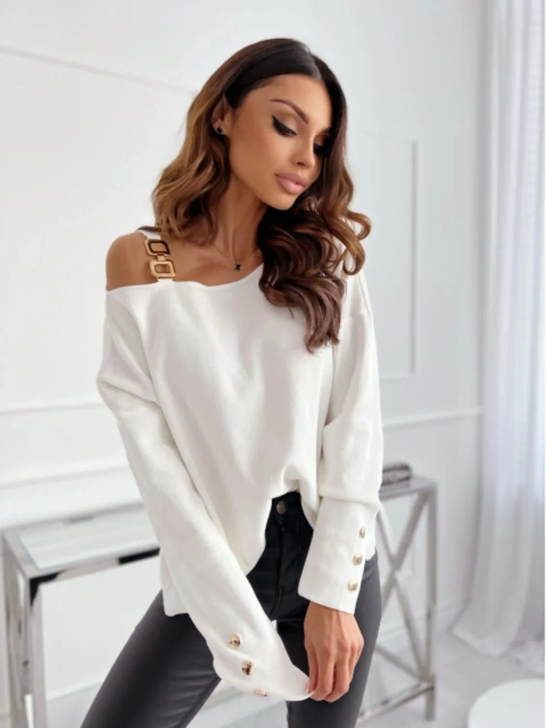 Woman wearing off-shoulder white sweater with gold buckle and black pants in modern room